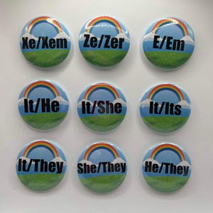 May include: Nine circular pin-back buttons arranged in a grid, each featuring a blue sky, a green grassy hill, white clouds, and a colorful rainbow. Each button displays different black text pronouns: Xe/Xem, Ze/Zer, E/Em, It/He, It/She, It/Its, It/They, She/They, and He/They.