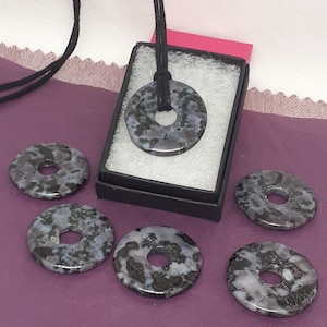 May include: Six black and white speckled donut-shaped pendants with a hole in the center. One pendant is on a black cord necklace in a small black box with a white interior. The pendants are on a purple surface.