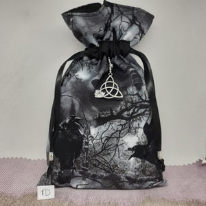 May include: Black drawstring bag with a silver Celtic knot charm. The bag is made of fabric with a black and white print of trees and ravens. The bag is tied with a black ribbon.