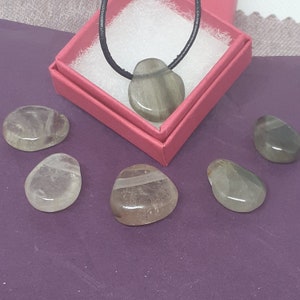 May include: A set of six smooth, polished stones in shades of gray and white. The stones are arranged on a purple surface. One stone is on a black cord and is in a red box.