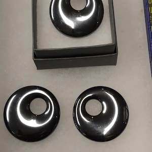 Puede incluir: Una colección de colgantes de hematita gris oscuro pulidos. Un colgante se muestra en un cordón negro dentro de una pequeña caja gris. Varios otros colgantes circulares con un agujero central están dispuestos debajo.