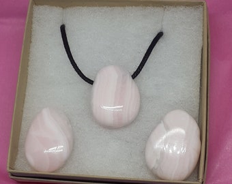May include: A set of pink and white stone jewelry, including a pendant on a black cord and two matching stones. The stones are displayed in a small, open cardboard box with white padding, set against a pink background.