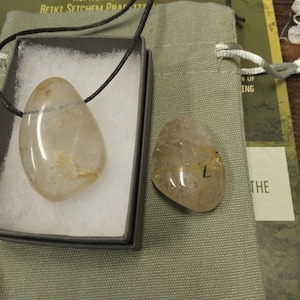 May include: A clear, polished quartz pendant with a black cord, displayed in a black box with white padding. A second, similar quartz stone with the letter "L" is next to a green drawstring bag. The stones have golden inclusions.