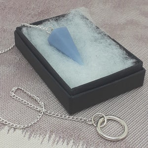 Puede incluir: Un péndulo de cristal azul claro con una cadena de plata, presentado en una caja negra con relleno blanco. El péndulo tiene forma de cono. La cadena está conectada a un anillo plateado.