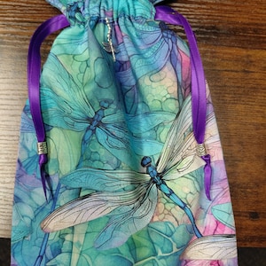 May include: A drawstring bag with a dragonfly pattern in shades of blue, teal, and purple. The bag features a repeating dragonfly design on a leaf-like background. It has purple ribbon drawstrings and a small dragonfly charm.