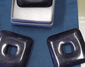 Könnte beinhalten: Drei quadratische Anhänger mit einem Loch in der Mitte. Die Anhänger sind aus schwarzem Stein mit glitzernder Oberfläche. Die Anhänger werden auf einem blauen Stoffhintergrund präsentiert.