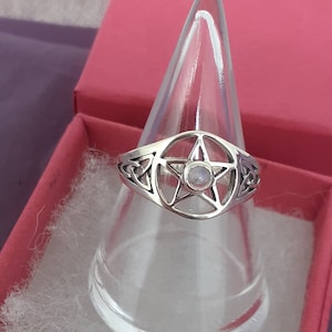 May include: A silver ring featuring a pentagram design with a clear stone at its centre. The ring is presented on a clear ring stand.