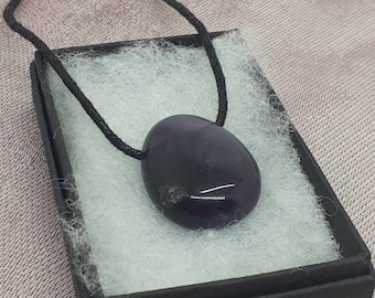 Puede incluir: Un collar de cordón negro con un colgante ovalado liso de color morado oscuro.