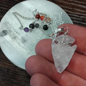 Op de afbeelding: Een heldere kwarts pijlpunthanger met een zilveren draadwikkel, vastgehouden in een hand. Een zilveren ketting met kleurrijke kralen is zichtbaar. De hanger ligt op een witte, geëtste plaat.