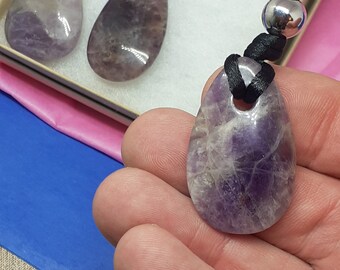 Puede incluir: Un collar con un colgante de amatista púrpura, un cordón negro y una cuenta de plata. El colgante tiene forma de lágrima y tiene una textura natural y rugosa.