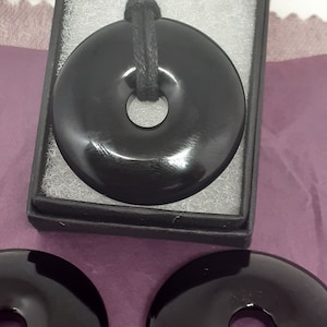 May include: A black obsidian donut pendant with a black cord. The pendant is in a black box with white lining. There are two more black obsidian donut pendants on a purple surface.