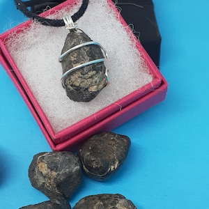 Pode incluir: Um pingente de pedra cinza escuro envolto em um fio de prata, pendurado em um cordão preto. O pingente está em uma caixa rosa com forro branco. Existem quatro pedras cinza escuras adicionais em uma superfície azul.