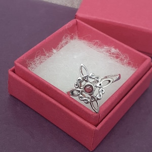May include: A silver ring with a pink gemstone in the center, set in a Celtic knot design. The ring is displayed in a red velvet ring box.
