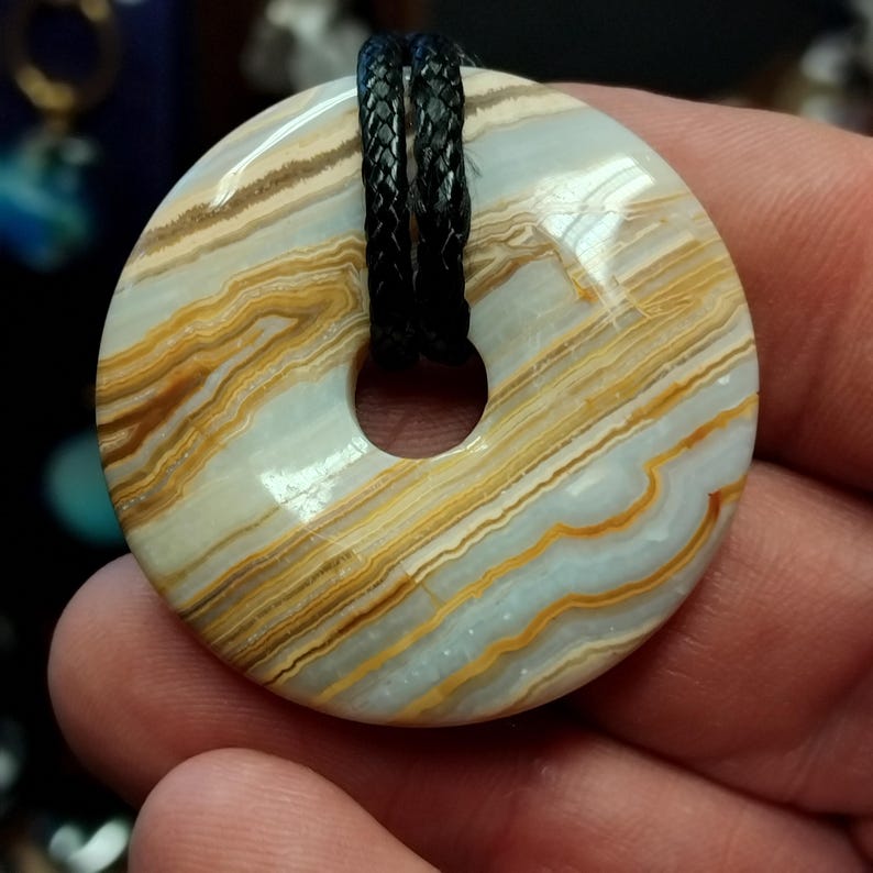 May include: A round, polished stone pendant with a central hole, featuring bands of tan, gold, and light blue. A black braided cord is threaded through the hole. The stone is approximately 2 inches in diameter.
