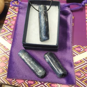 May include: A blue kyanite pendant on a black cord, presented in a small black box with white padding. Two additional kyanite stones are beside the box on a purple velvet pouch. The pouch is on a patterned surface.