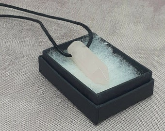Puede incluir: Un colgante de cristal transparente rosa pálido en un cordón negro, presentado en una caja de regalo negra con papel de seda blanco.