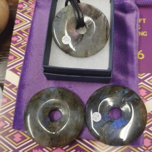 May include: Three gray and blue labradorite donut pendants. One is displayed in a black box with a black cord. Two additional pendants are displayed below. Each pendant has a hole in the center and a small white sticker.