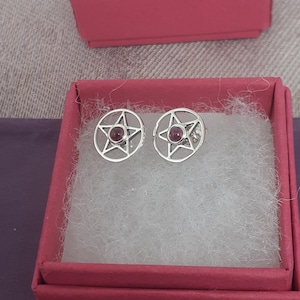 Op de afbeelding: Een paar zilveren oorbellen met een pentagram-ontwerp en een rode edelsteen in het midden van elk.