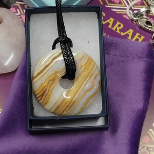 May include: A circular pendant necklace with a natural stone donut-shaped pendant. The stone features bands of white, tan, and gold. The pendant is suspended from a black cord and presented in a blue box.