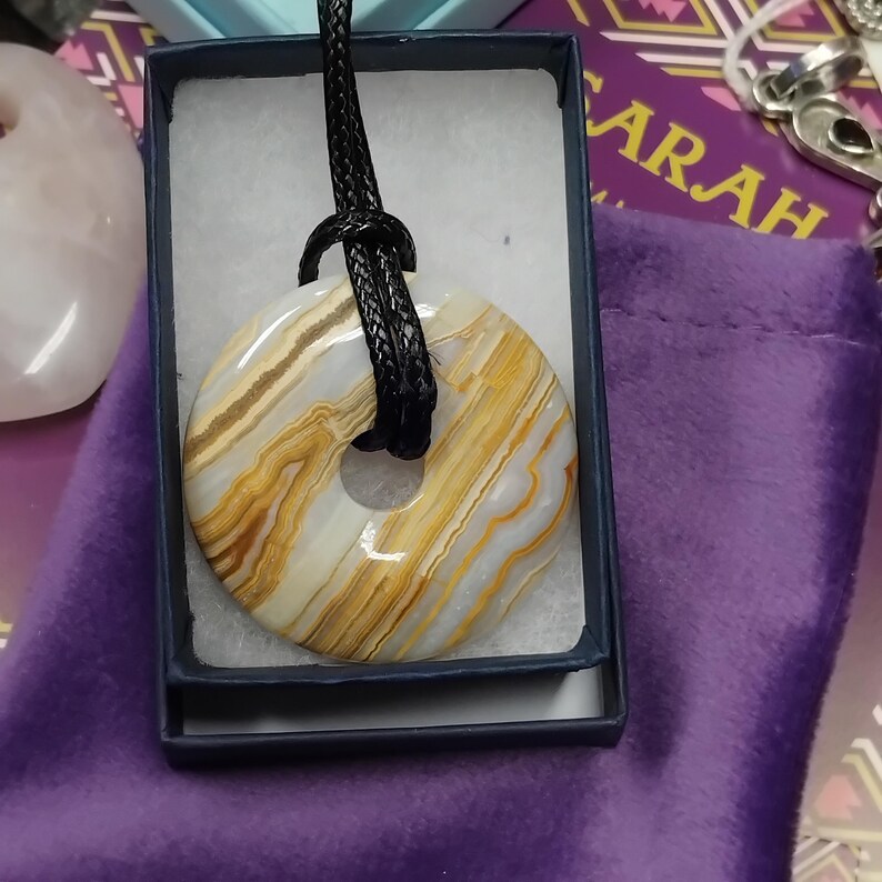 May include: A circular pendant necklace with a natural stone donut-shaped pendant. The stone features bands of white, tan, and gold. The pendant is suspended from a black cord and presented in a blue box.