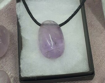 Puede incluir: Un colgante de amatista púrpura en un collar de cordón negro. El colgante está en una pequeña caja con forro blanco.