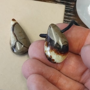 Può includere: Due ciondoli a forma di lacrima realizzati in agata a bande marroni, bianche e nere. I ciondoli sono su un cordoncino nero.