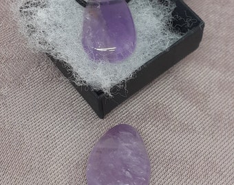 Puede incluir: Dos piedras de amatista, una en un collar de cordón negro y otra suelta, ambas con un tono púrpura claro y un acabado liso y pulido.