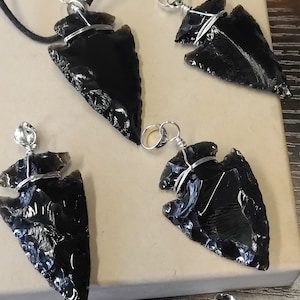 Puede incluir: Cuatro colgantes de punta de flecha de obsidiana negra con alambre plateado envuelto alrededor de la parte superior. Las puntas de flecha son todas de diferentes formas y tamaños.