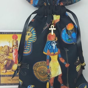 Puede incluir: Bolsa de tela negra con un amuleto Ankh dorado. La bolsa está decorada con un tema egipcio, incluyendo un faraón, un escarabajo y un ojo de Horus. La bolsa está atada con una cinta negra y tiene cuentas doradas en los extremos de las cintas. La bolsa está sentada sobre una superficie blanca junto a una carta de tarot con el número VIII y la palabra FUERZA.