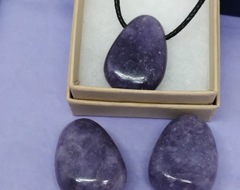 Puede incluir: Tres piedras de lepidolita púrpura, dos son lisas y una está en un collar de cordón negro. Las piedras están en una caja blanca con una tapa azul.