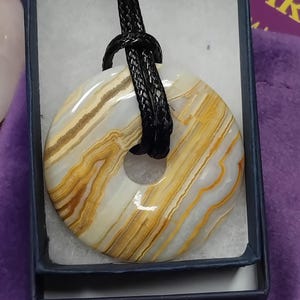 May include: A donut-shaped pendant with a black cord. The pendant is made of a stone with layers of white, tan, and gold colors. The pendant is displayed in a small, dark blue box with a white interior.