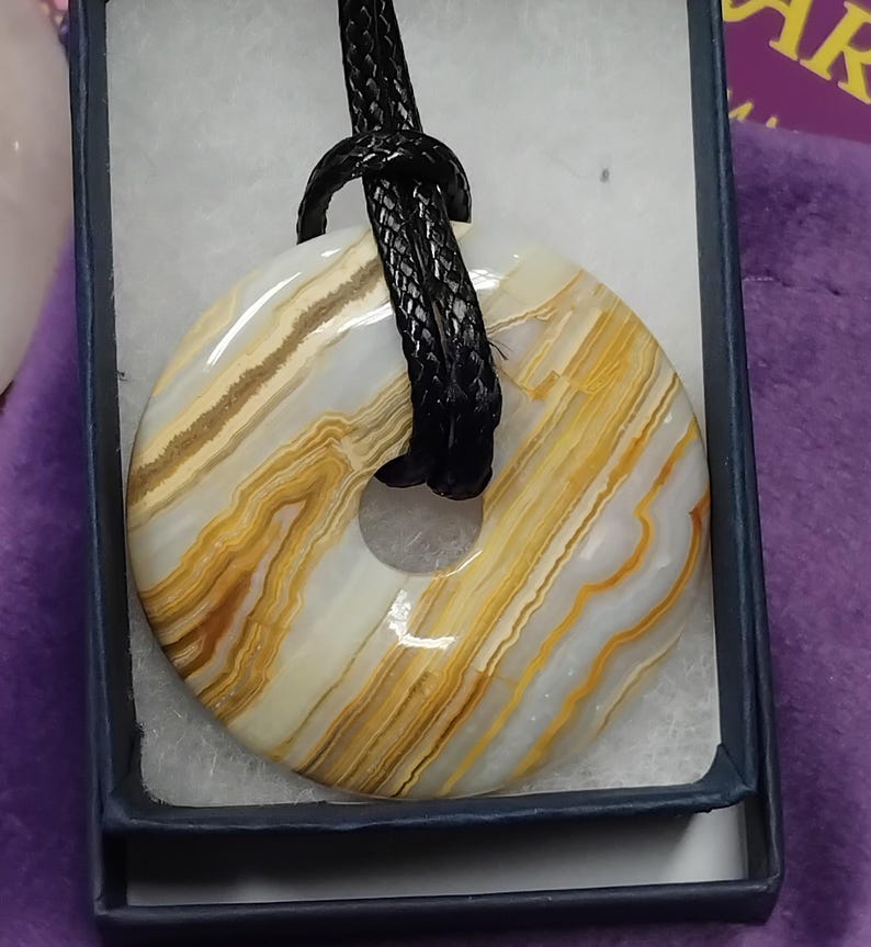 May include: A donut-shaped pendant with a black cord. The pendant is made of a stone with layers of white, tan, and gold colors. The pendant is displayed in a small, dark blue box with a white interior.