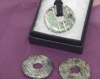 Puede incluir: Un collar con un colgante redondo y plano de piedra verde y blanca. El colgante tiene un patrón de remolino y está en un cordón negro.