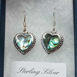 May include: A pair of sterling silver heart-shaped earrings with abalone shell inlays. The earrings are in a blue jewelry box with white padding.