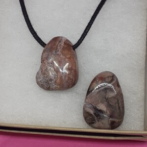 May include: Two brown and white agate stones on a black cord necklace. The stones are irregularly shaped and have a natural, polished finish.