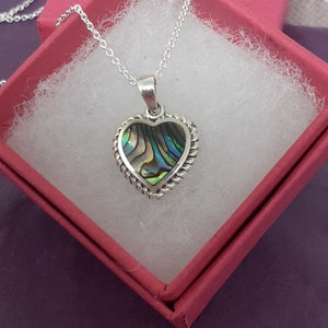 Puede incluir: Un collar de plata con un colgante en forma de corazón con una incrustación de concha de abulón verde, azul y morado. El collar está en una caja de regalo roja.