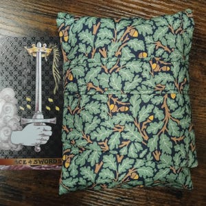 Puede incluir: Una bolsa de tela verde y azul marino con un diseño de hojas de roble y bellotas. Junto a la bolsa, una carta de tarot con una mano sosteniendo una espada y el texto "ACE of SWORDS".