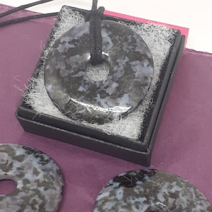 May include: A black and blue stone pendant with a hole in the center, hanging from a black cord. The pendant is in a small black box with white lining.