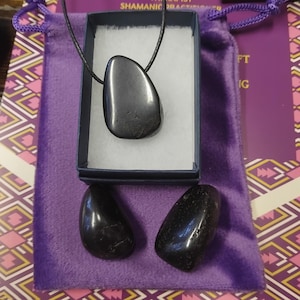 Pode incluir: Um pingente de pedra cinza escuro polida em um cordão preto, apresentado em uma pequena caixa azul com interior branco. Duas pedras polidas adicionais estão localizadas abaixo da caixa em uma bolsa de veludo roxo. A bolsa está em uma superfície estampada.