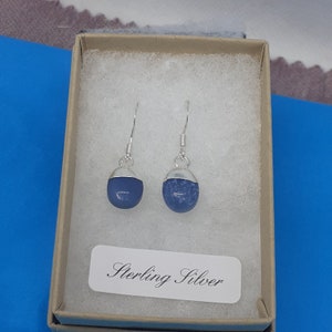 May include: A pair of sterling silver earrings with blue stone cabochons. The earrings are presented in a small, open cardboard box with a white label that reads "Sterling Silver". The earrings have a simple, elegant design.