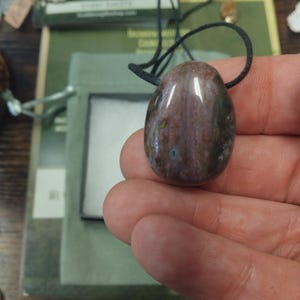 Könnte beinhalten: Ein ovaler, polierter Anhänger mit einer schwarzen Kordel. Der Anhänger hat eine Mischung aus braunen, grünen und weißen Farben. Der Stein ist glatt und reflektierend, mit einem natürlichen Muster. Der Anhänger wird in einer Hand gehalten.