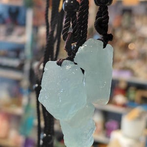 May include: A necklace featuring three raw, pale blue, translucent crystal pendants suspended from dark brown twisted cord strings. Each crystal has an irregular, jagged shape with a textured surface. Small, dark, spherical beads are threaded onto the cords.