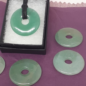 May include: Four green jade donut pendants, one of which is on a black cord necklace in a black box. The pendants are smooth and have a hole in the center.
