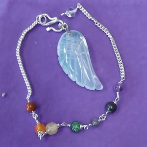 Puede incluir: Un brazalete de cadena de plata con un dije de ala de ángel blanco y cuentas de piedras preciosas de colores. Las cuentas son rojas, naranjas, amarillas, verdes, grises y moradas.