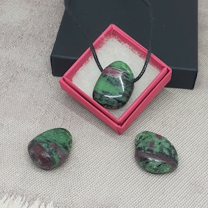 Könnte beinhalten: Drei grüne und rosafarbene Edelstein-Anhänger, einer an einer schwarzen Schnurkette, in einer rosa Schachtel. Die Edelsteine sind glatt und oval geformt.