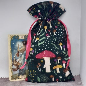 May include: A green drawstring bag with a mushroom and leaf print. The bag has a pink drawstring and is closed. The bag is sitting on a white surface. The bag is decorated with a variety of mushrooms, including red, white, and brown mushrooms.