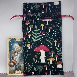 May include: A green fabric pouch with a drawstring closure. The pouch is decorated with a repeating pattern of white mushrooms and green leaves. The pouch is ideal for storing small items like tarot cards or jewellery.