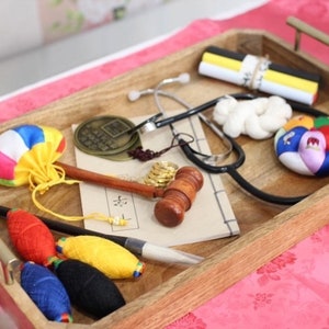 May include: A wooden tray holds various items, including a stethoscope, a small gavel, a coin, a colorful pouch, and spools of thread. The tray has brass handles and sits on a pink floral surface.