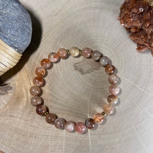 May include: A beaded bracelet featuring round, polished stones in shades of orange, brown, and cream. The bracelet is strung with a gold-colored elastic cord and has small gold-colored beads interspersed throughout. The bracelet is displayed on a wooden surface.