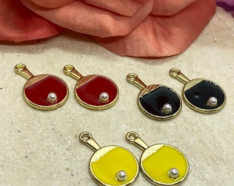 May include: Six gold-tone table tennis paddle charms with a white pearl in the center of each paddle. Two charms are red, two are black, and two are yellow.